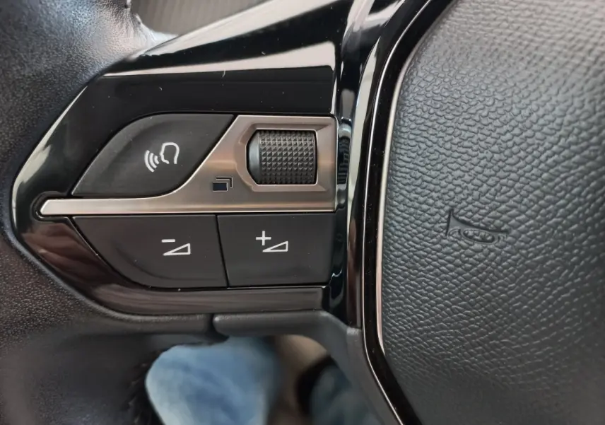 Gros plan sur les commandes de volume et reconnaissance vocale sur le volant cuir noir d'une Peugeot 208 blanche.