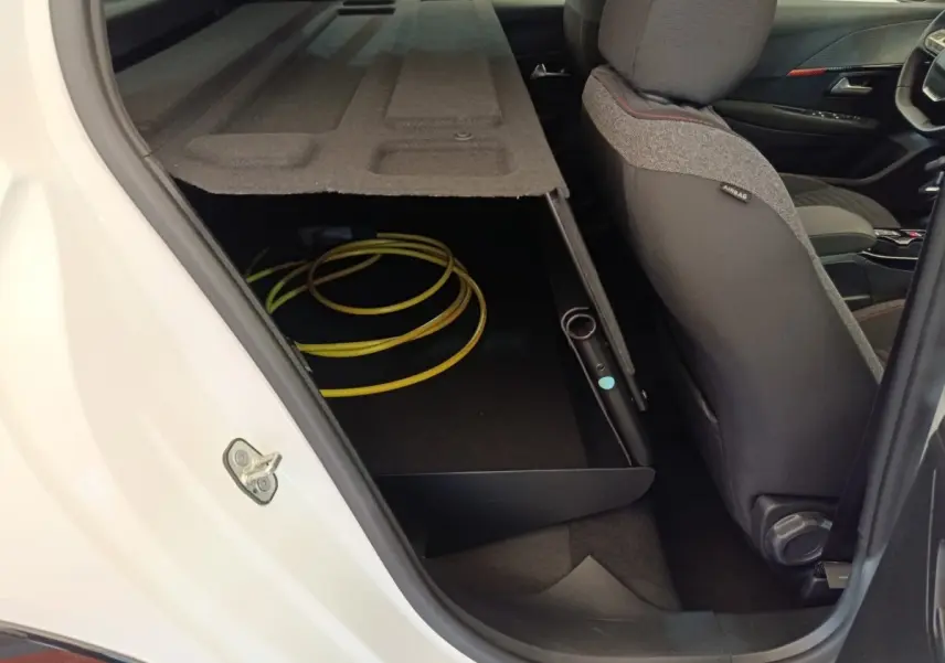 Vue intérieure côté arrière droit d'une Peugeot 208 électrique blanche, montrant le rangement sous la banquette avec câble de charge.