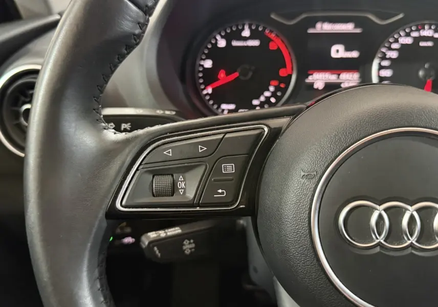 Gros plan sur le volant cuir noir d'Audi A3 2.0 TDI 2018 avec commandes multifonctions et compteurs en arrière-plan flous.
