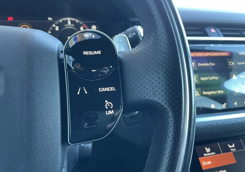 Gros plan sur les commandes de régulateur de vitesse au volant cuir perforé du Range Rover Velar gris Corris, tableau de bord numérique visible.