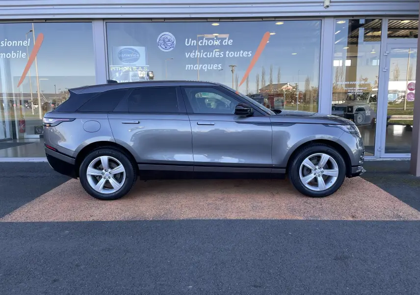 Profil côté gauche d'un Range Rover Velar D240 S R-Dynamic gris Corris, avec jantes alliage et toit noir devant un showroom.