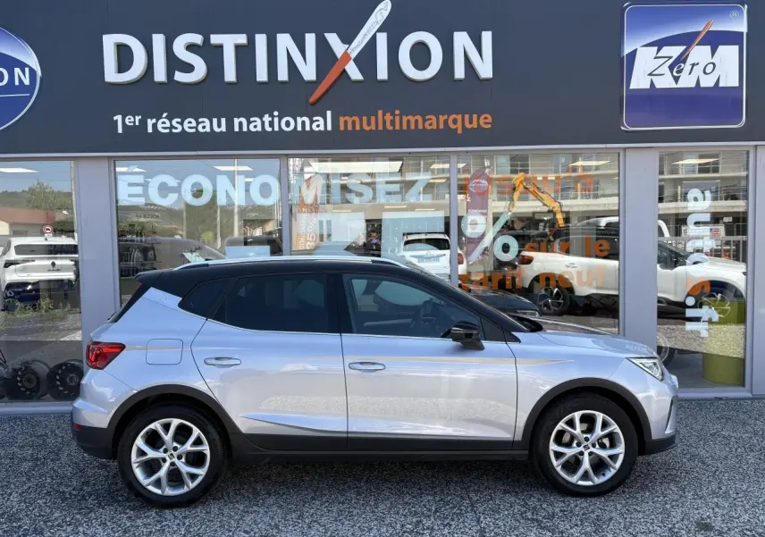 Vue de profil droit d’un SEAT Arona 2024 gris clair avec toit noir et jantes alliage 16 pouces devant un showroom Distinxion.