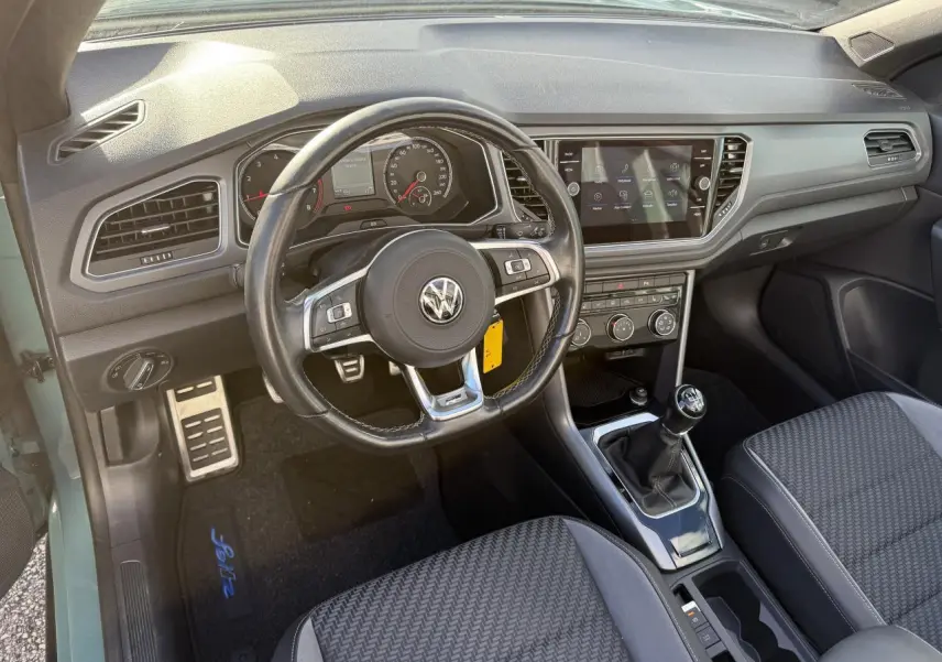 Vue intérieure côté conducteur du Volkswagen T-Roc Cabriolet vert cactus, volant cuir multifonction et boîte manuelle visible.