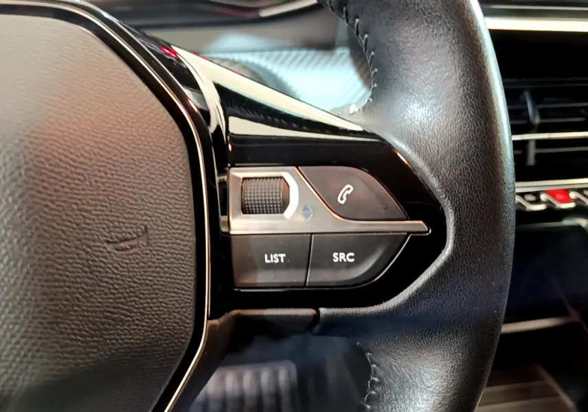 Gros plan sur les commandes gauche du volant cuir noir du Peugeot 2008 gris clair, avec tableau de bord en arrière-plan.