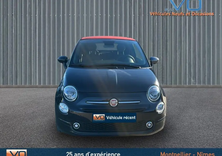 Vue frontale d'une FIAT 500C noire avec toit rouge, modèle 2024, affichée sur un parking devant un mur métallique.