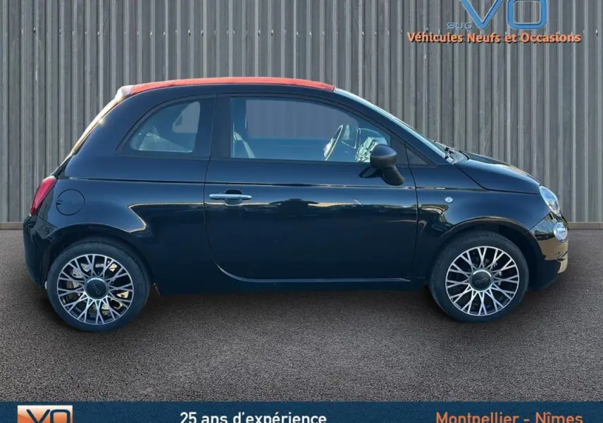 Profil côté gauche d'une FIAT 500C noire 2024 avec toit rouge et jantes stylisées, stationnée devant un mur métallique.