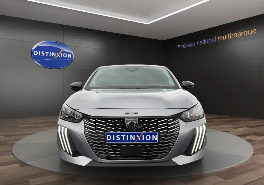 Vue frontale d'une Peugeot 208 gris clair avec calandre noire et feux LED verticaux allumés en showroom.