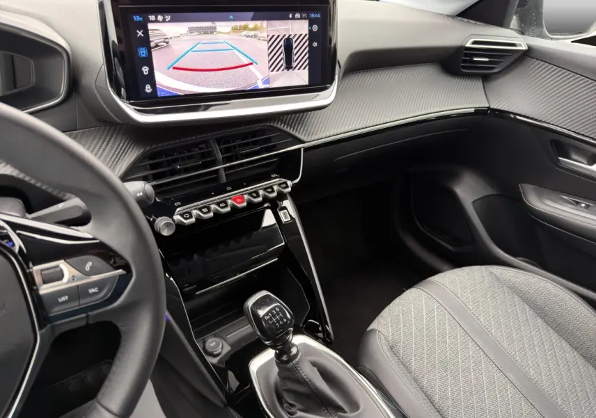 Vue intérieure côté conducteur de la Peugeot 208 gris clair, avec écran de caméra de recul et levier de vitesse manuel visible.