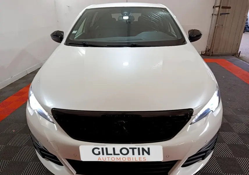 Vue avant d'une Peugeot 308 blanche avec calandre noire et phares LED allumés dans un garage.