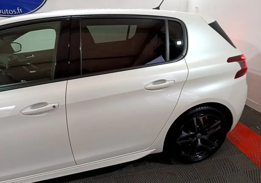 Profil côté gauche d'une Peugeot 308 blanche avec jantes noires et vitres teintées en intérieur showroom.