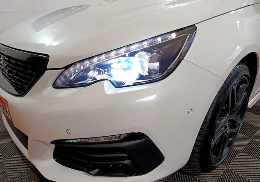Gros plan sur l'avant droit blanc d'une Peugeot 308 2019 avec phare LED allumé et jante noire brillante