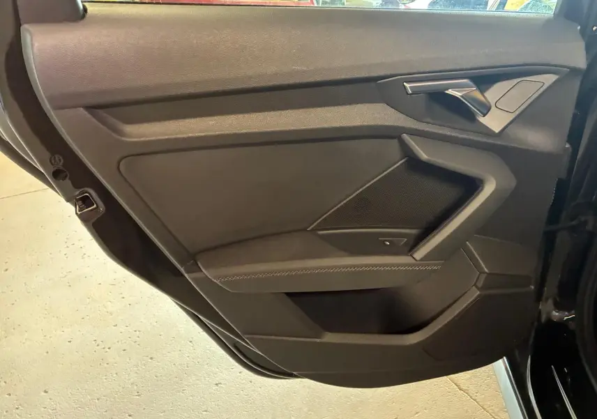 Vue intérieure côté gauche de la porte arrière noire de l'Audi A3 Sportback avec accoudoir en similicuir et poignée argentée.