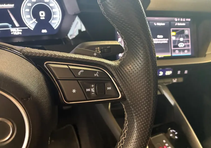 Gros plan sur le volant cuir perforé côté gauche et tableau de bord numérique de l'Audi A3 Sportback noir 2023.