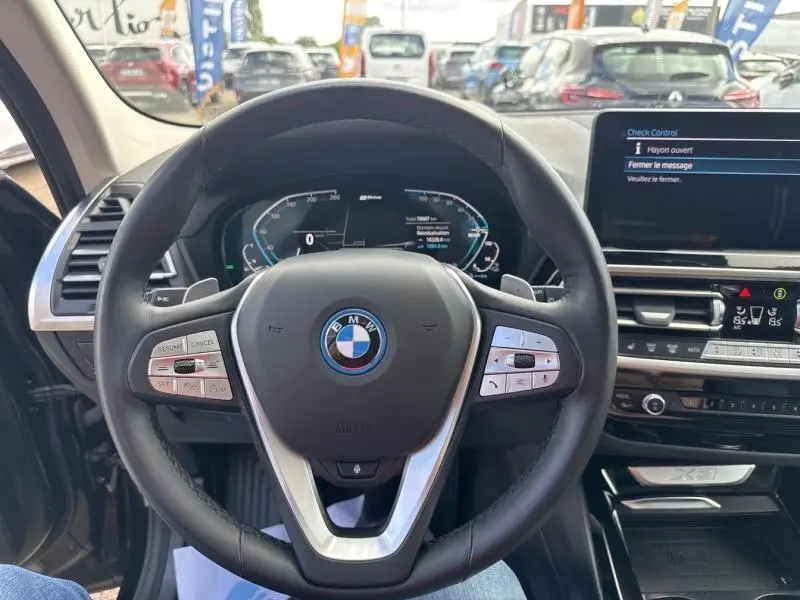 Vue intérieure centrée sur le volant cuir multifonction d'une BMW X3 xDrive30e 2023 avec tableau de bord numérique et écran tactile.