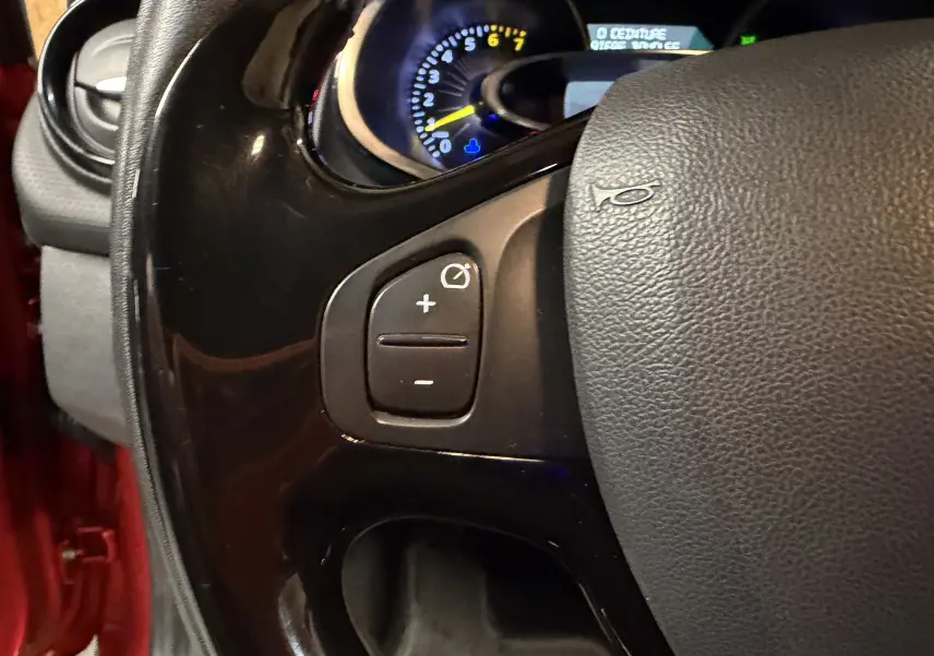 Gros plan sur le volant noir de la Renault Clio Estate IV rouge, montrant les commandes de régulateur de vitesse côté gauche.