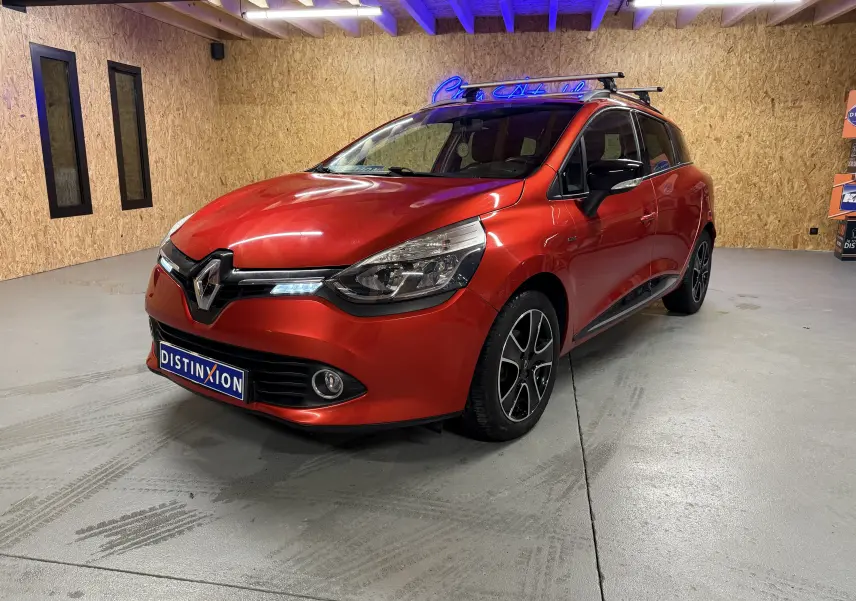 Renault Clio Estate IV rouge vue 3/4 avant droit, avec jantes noires et barres de toit en intérieur showroom.