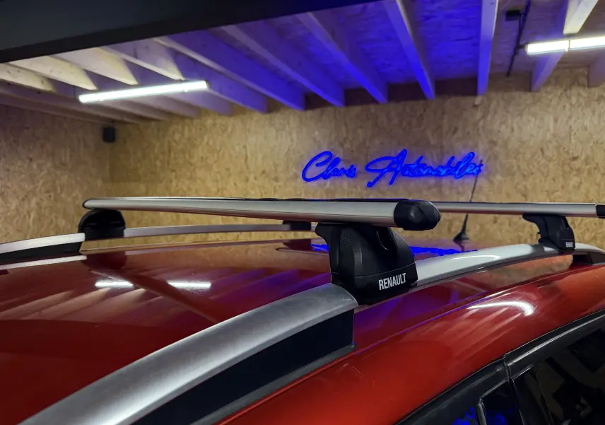 Barres de toit Renault sur le toit rouge d'une Clio Estate IV, vue rapprochée en intérieur avec éclairage bleu.