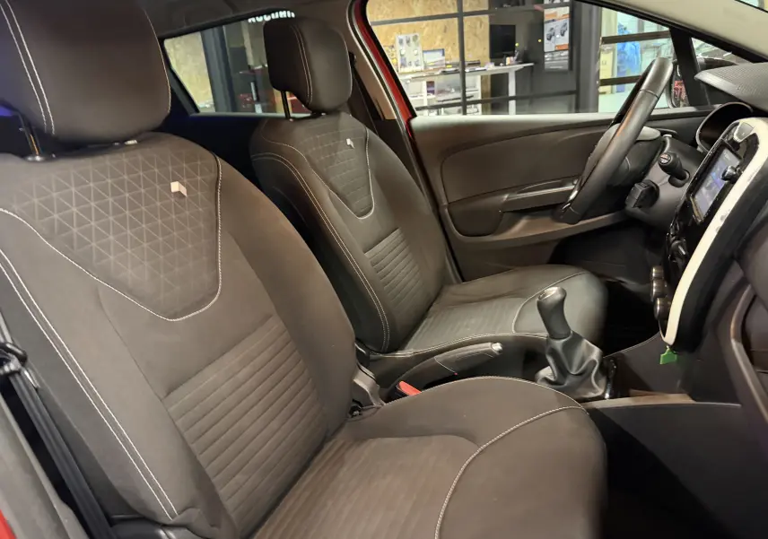 Vue intérieure côté conducteur de la Renault Clio Estate IV rouge, montrant sièges noirs et levier de vitesse manuel.