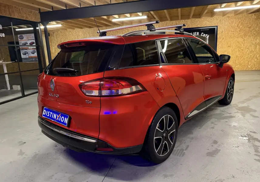 Vue 3/4 arrière droite d'une Renault Clio Estate IV rouge avec barres de toit et attelage, stationnée en intérieur.