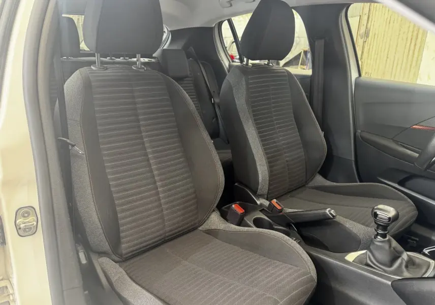 Vue intérieure côté avant droit de la Peugeot 208 blanche, montrant les sièges tissu noirs et la boîte manuelle.