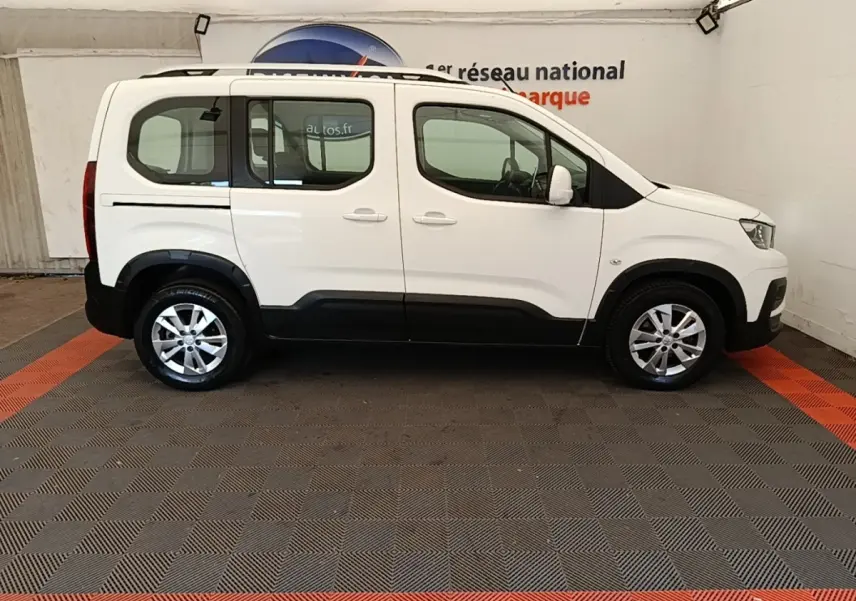 Profil droit d’un Peugeot Rifter blanc 2020 avec barres de toit chromées et jantes alliage en intérieur showroom.