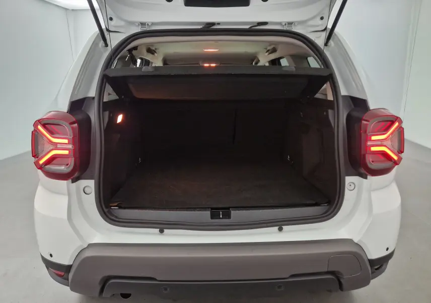 Coffre ouvert vu de l'arrière d'un Dacia Duster blanc 2024, feux arrière allumés et intérieur du coffre spacieux visible.