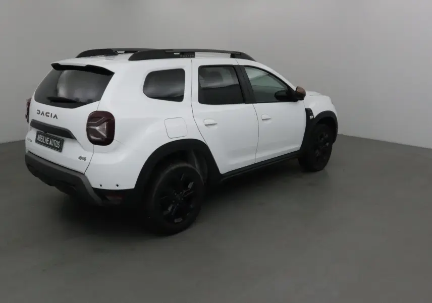 Vue 3/4 arrière droite d'un Dacia Duster 4x4 blanc 2024 avec jantes noires et barres de toit noires.