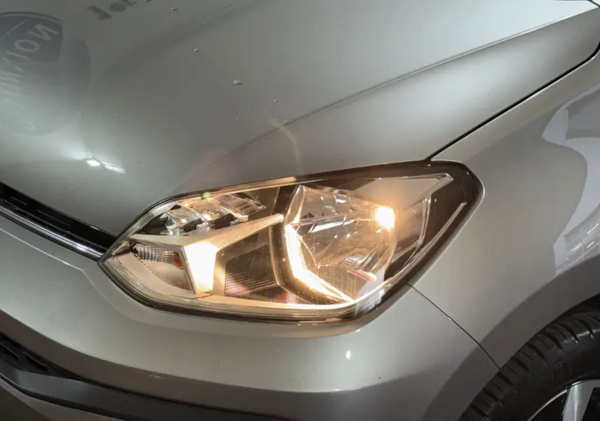 Gros plan sur le phare avant droit allumé d'une Volkswagen Up gris foncé, avec partie du capot et aile visible.
