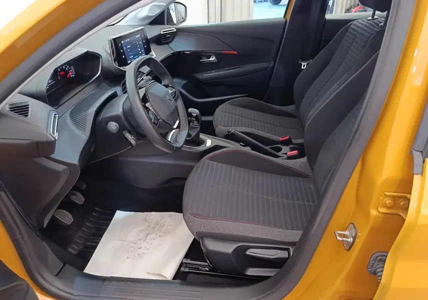 Vue intérieure côté conducteur de la Peugeot 208 jaune 2023, volant cuir multifonctions et sièges tissu gris à surpiqûres rouges.