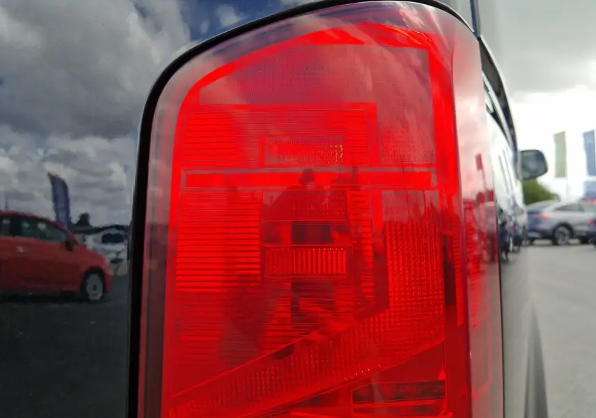 Gros plan sur le feu arrière droit rouge et orange d'un Volkswagen Transporter noir, reflet du ciel et véhicules alentours visible.