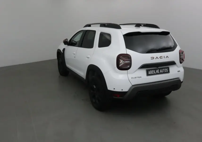 Vue 3/4 arrière droite d'un Dacia Duster blanc 2024 avec jantes noires et barres de toit noires.