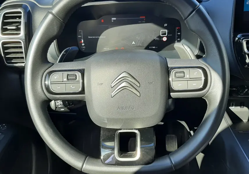 Vue rapprochée du volant noir du Citroën C5 Aircross 2024, avec commandes intégrées et tableau de bord numérique visible.