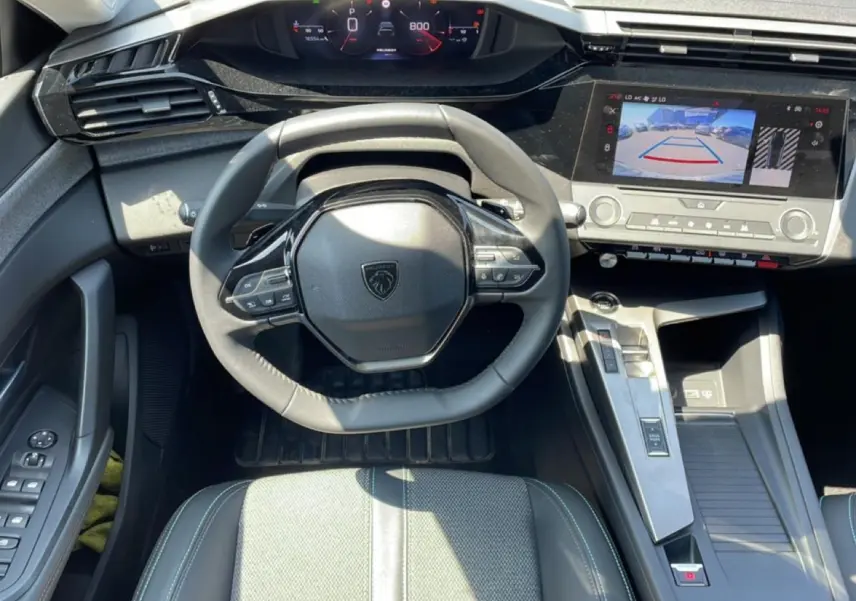 Vue intérieure avant centrée sur le volant cuir noir et l'écran tactile affichant la caméra de recul d'une Peugeot 308 gris 2025.