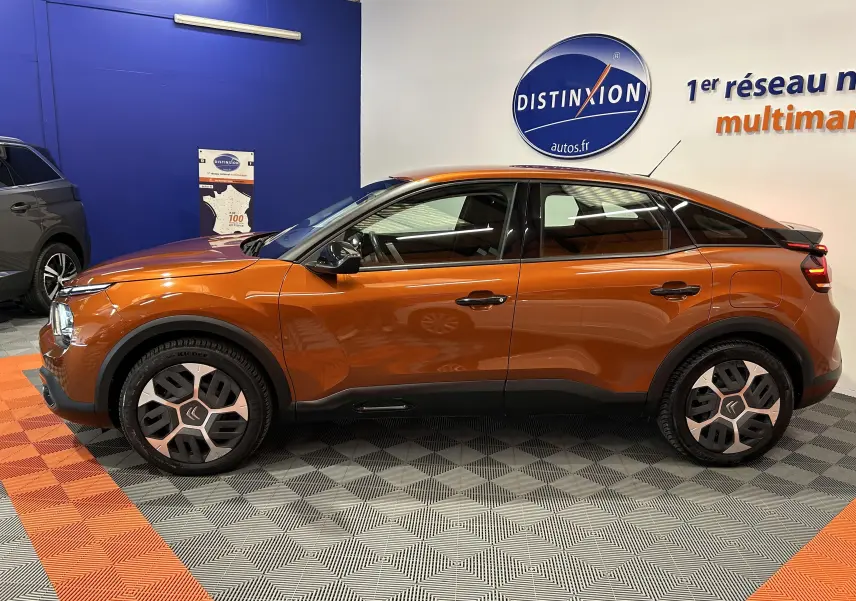 Profil côté gauche d'une Citroën C4 PureTech 100 Feel orange, avec jantes distinctives et attelage visible en intérieur showroom.