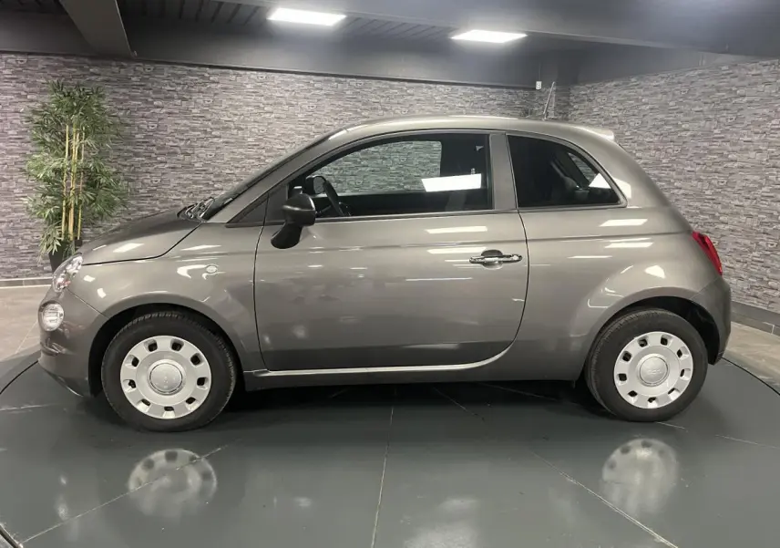 Profil droit d'une Fiat 500 gris métallisé 2023 avec jantes acier et rétroviseurs noirs dans un showroom.