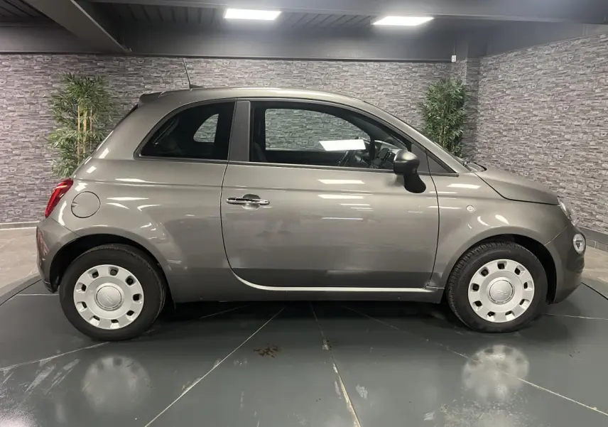Profil droit d'une Fiat 500 gris métallisé 2023 avec jantes acier et poignées chromées en intérieur showroom.
