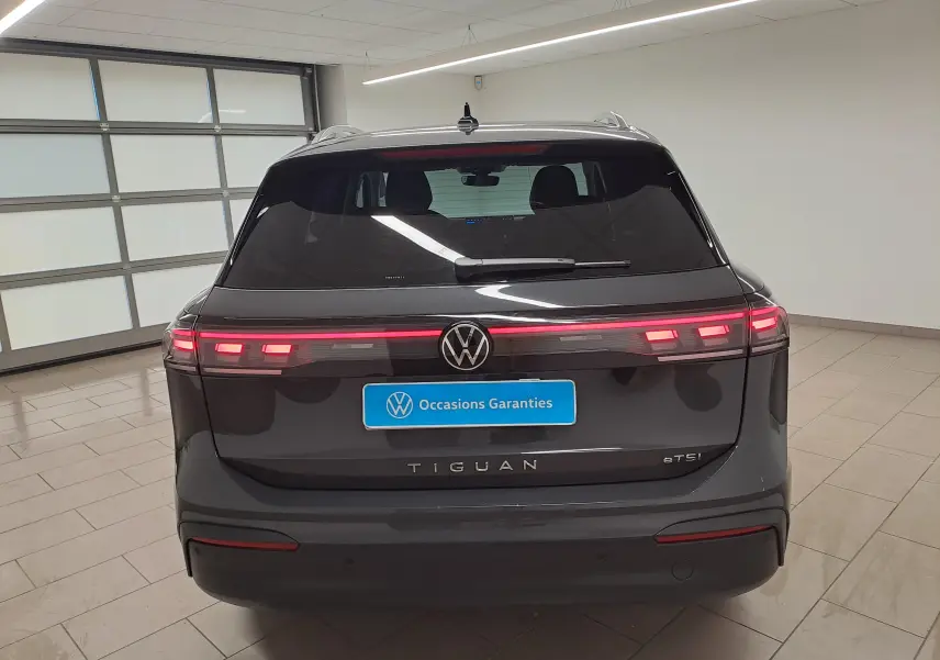 Vue arrière d'un Volkswagen Tiguan gris Urano 2024 avec feux allumés et plaque "Occasions Garantis" en intérieur showroom.