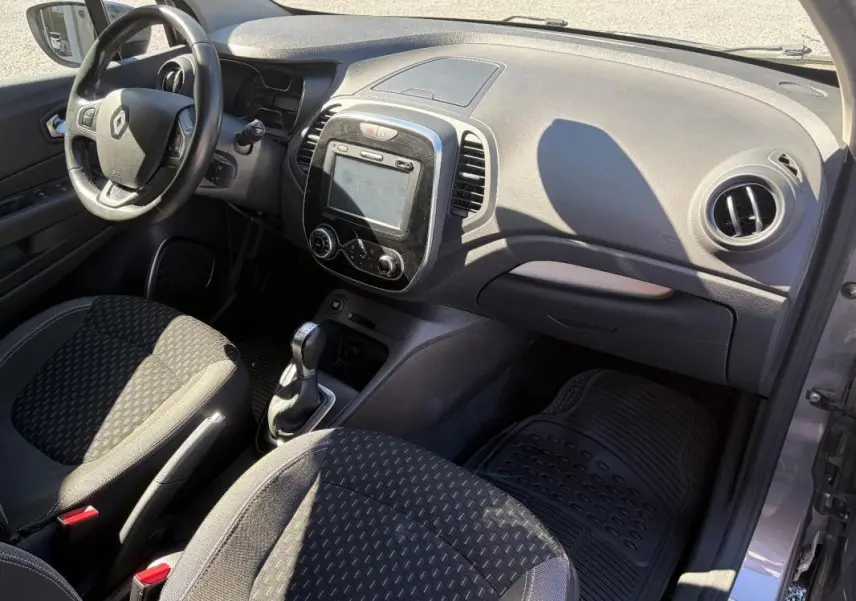 Vue intérieure côté conducteur du Renault Captur gris, montrant le volant multifonctions, la console centrale avec écran tactile et la boîte automatique.