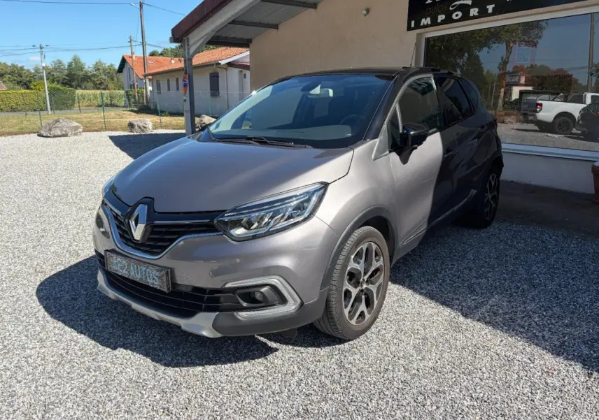 Renault Captur gris 3/4 avant droit, avec jantes alliage et calandre noire brillante sous un soleil clair.