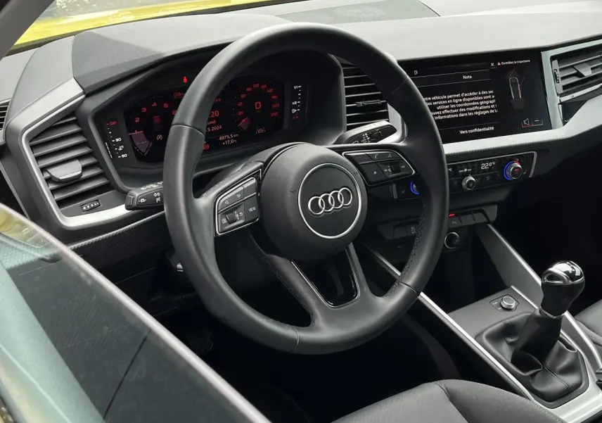 Vue intérieure côté conducteur de l'Audi A1 Allstreet jaune Python, volant cuir multifonctions et tableau de bord digital visibles.