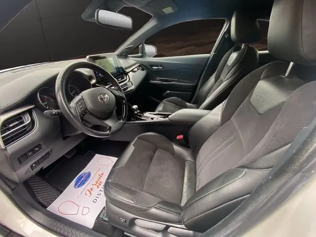 Vue intérieure côté conducteur du Toyota C-HR Hybride 2018 avec sièges noirs en cuir et tissu, tableau de bord moderne et écran tactile.