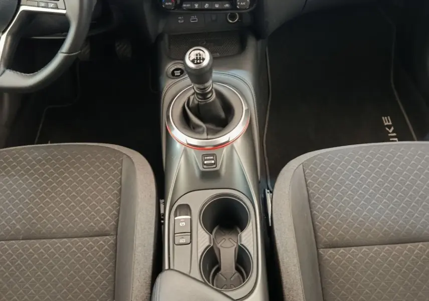 Vue intérieure centrée sur la console centrale et levier de vitesse manuel du Nissan Juke gris clair, avec accoudoir et sièges noirs.