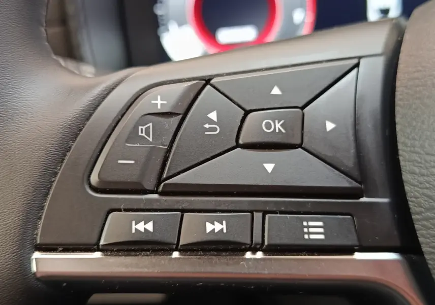 Gros plan sur les commandes multifonctions noires du volant du Nissan Juke 2025, avec affichage numérique flou en arrière-plan.