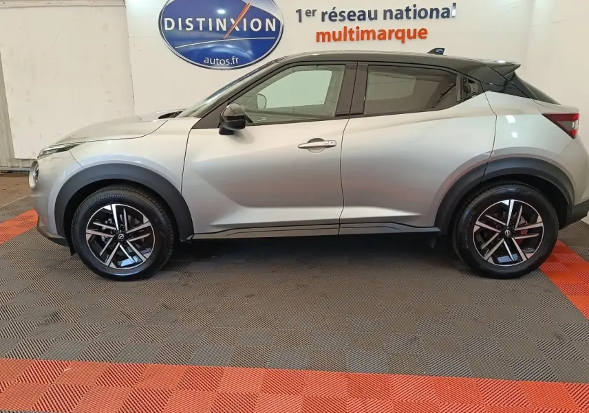 Vue de profil côté gauche d'un Nissan Juke gris clair 2025 avec toit noir et jantes alliage bicolores.