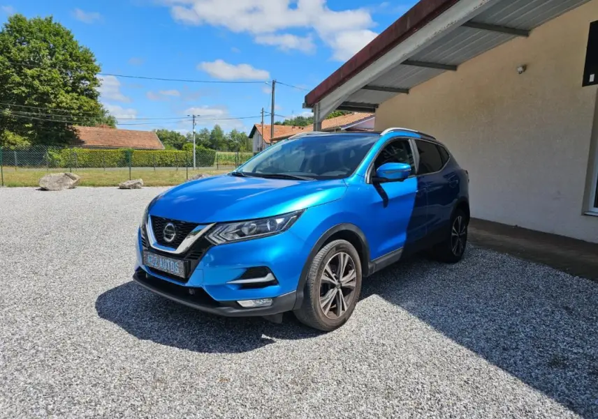 Nissan Qashqai bleu 2019 en 3/4 avant droit, stationné sur gravier devant un bâtiment beige.