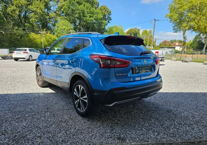 Nissan Qashqai bleu en 3/4 arrière droit, avec jantes alliage et toit ouvrant visible, stationné sur un sol gravillonné.