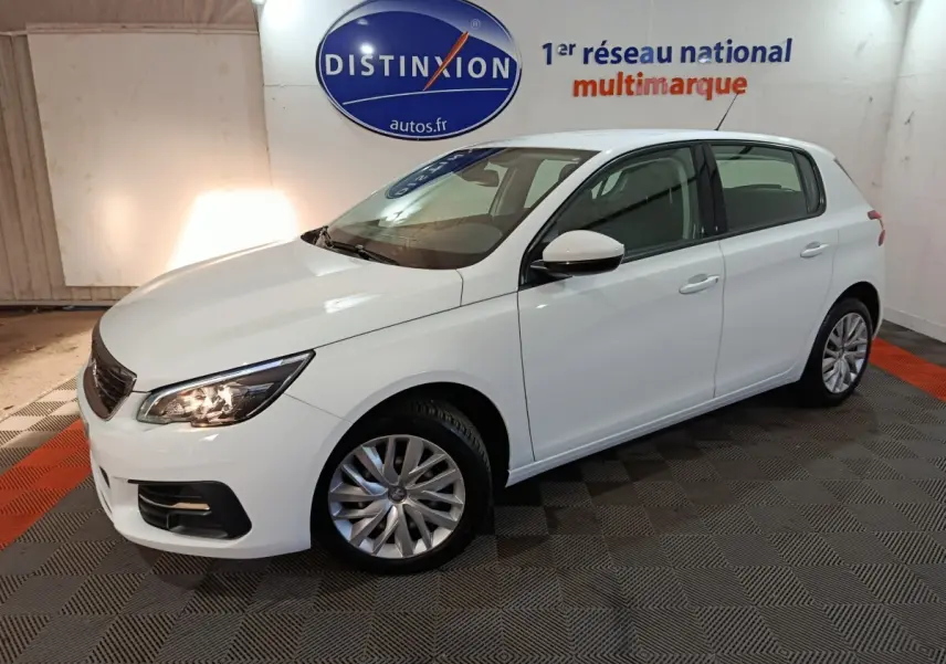 Peugeot 308 blanc vue 3/4 avant droit, phares allumés, stationnée en intérieur sur sol damier noir et rouge.