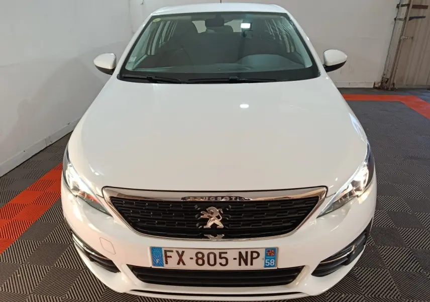 Vue avant d'une Peugeot 308 blanche 2021 avec calandre noire et phares allumés dans un garage.