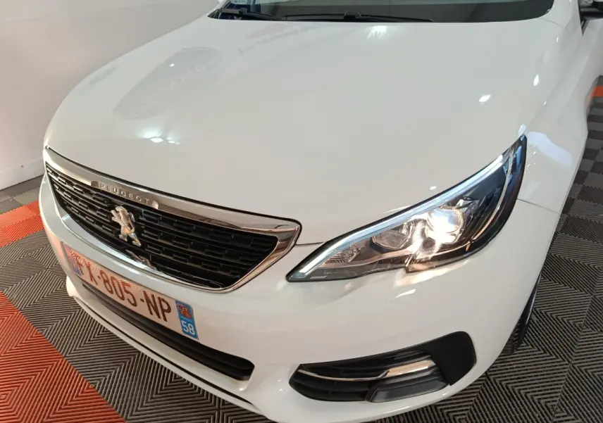 Vue 3/4 avant droit d'une Peugeot 308 blanche avec phares allumés et calandre chromée visible