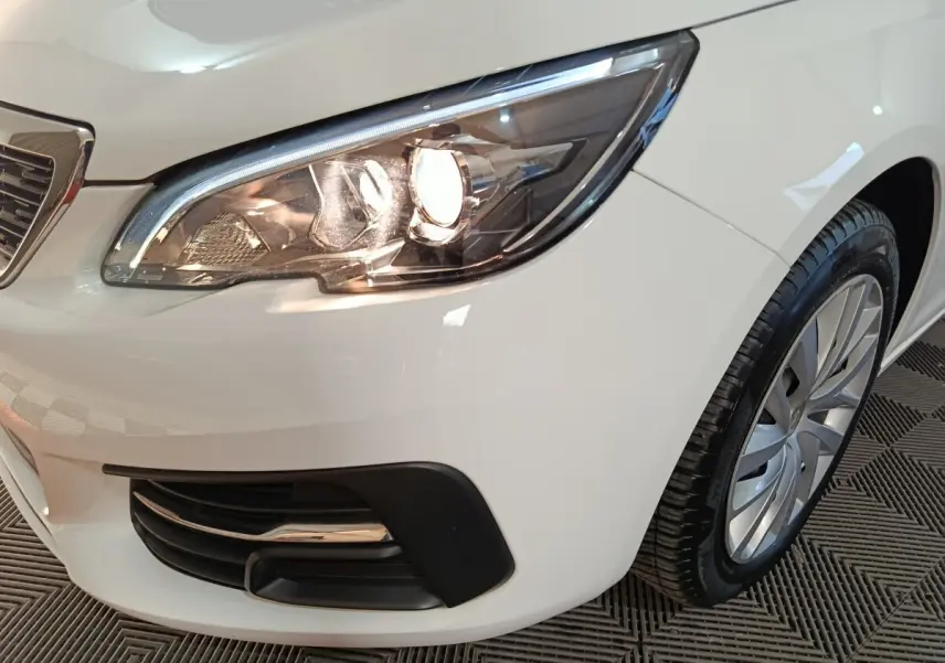 Gros plan sur le phare avant droit allumé d'une Peugeot 308 blanche, avec la roue et la calandre partiellement visibles.